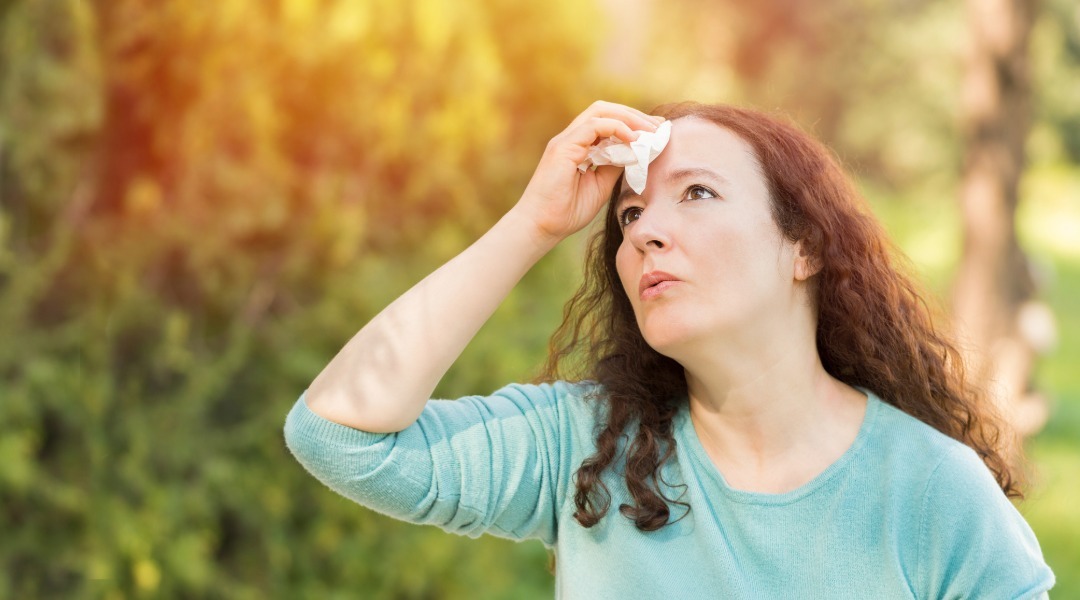 Woman outdoors on a sunny day wiping sweat from her forehead with a tissue, appearing overheated while surrounded by trees and greenery.