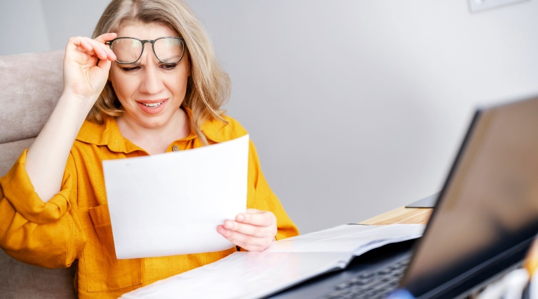 Woman wearing a yellow blouse looking confused while reviewing paperwork at a desk with a laptop, illustrating the challenges of understanding a drinking water quality report.