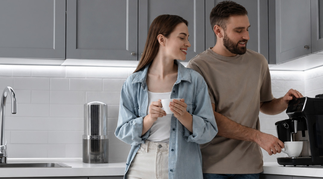 Couple enjoying coffee in a modern kitchen with gray cabinets and white countertops, with a Multipure Aquaperform drinking water filtration system visible on the counter behind them.