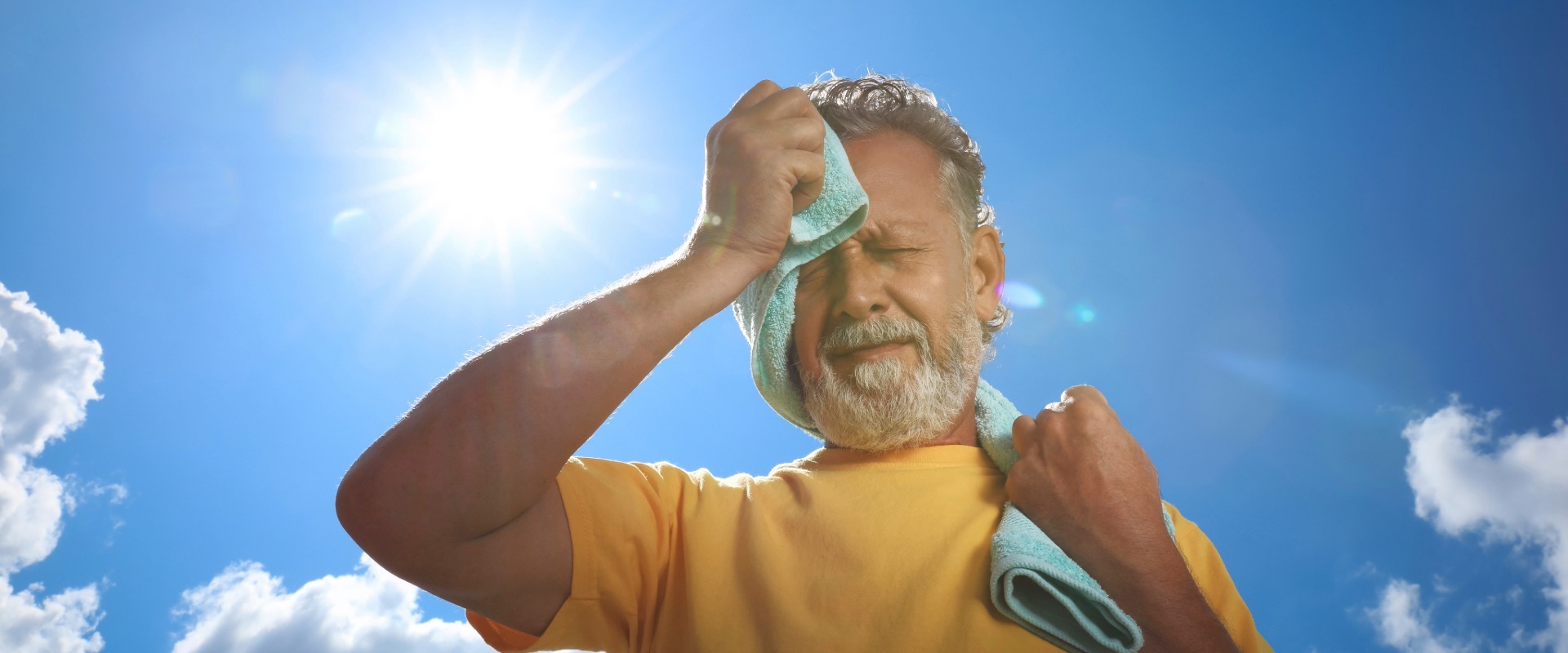 Older man in a yellow shirt wiping sweat from his forehead with a light blue towel while standing outdoors beneath a bright sun and clear blue sky.