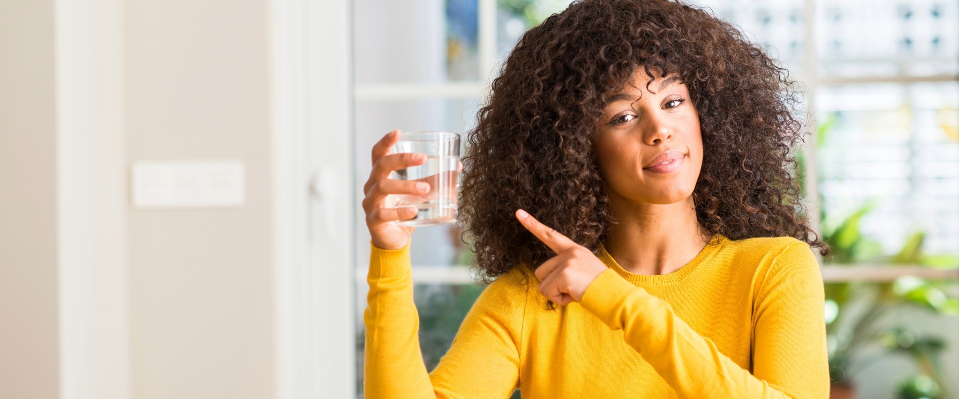 Woman in a yellow sweater holding a glass of water and pointing toward it while smiling in a bright, modern kitchen.