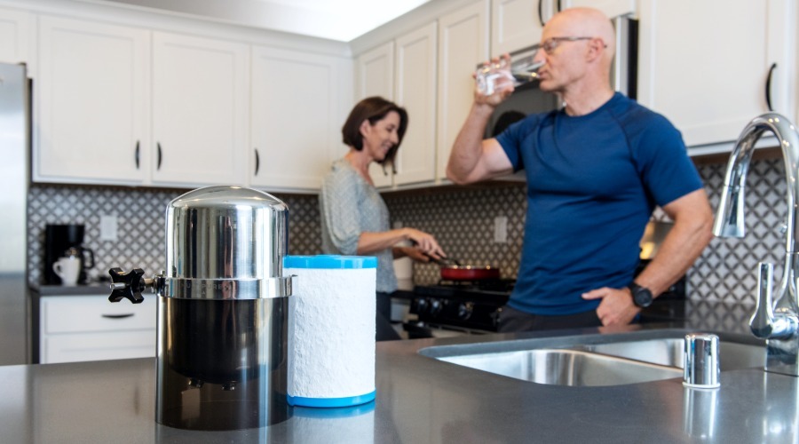 Multipure Aquaversa countertop water filtration system and replacement filter cartridge on a kitchen counter with homeowners in the background.