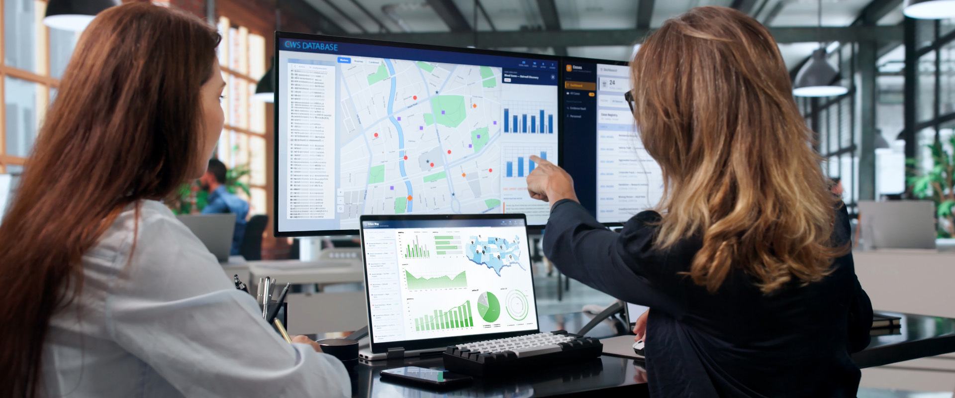 Two women analyzing geographic data and statistical charts on computer screens in a modern office, reviewing maps, graphs, and database information.